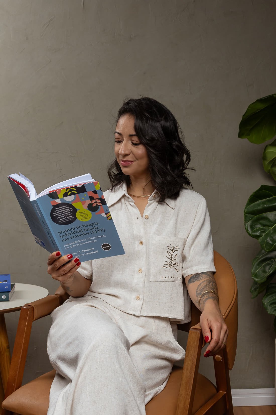 Karla Rocha sentada na poltrona do consultório, lendo um manual de psicoterapia